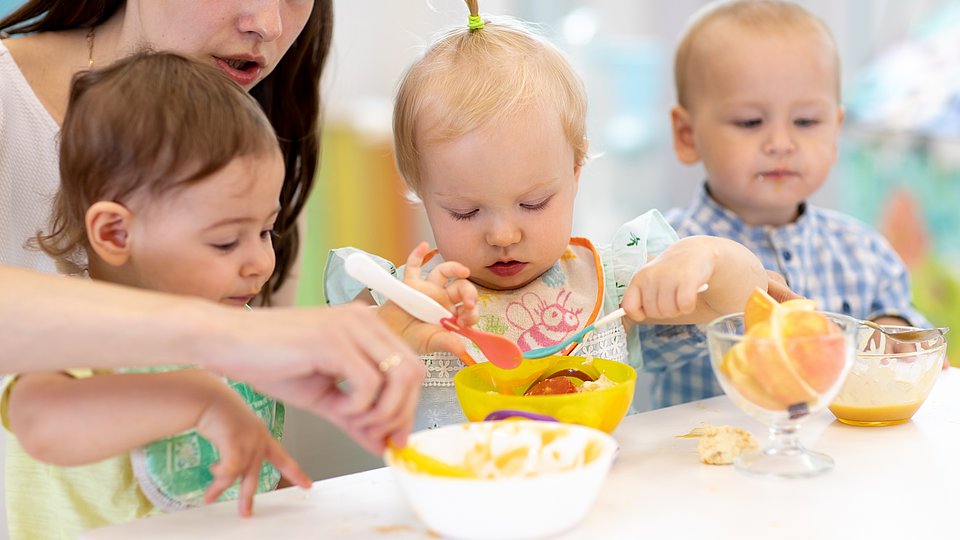 Erzieherin sitzt mit drei Kleinkindern am Tisch und hilft ihnen beim Essen.