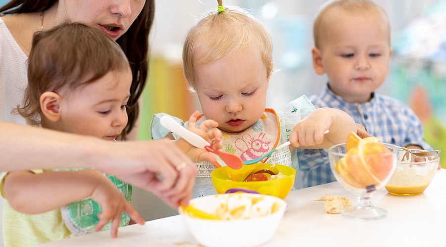 Erzieherin sitzt mit drei Kleinkindern am Tisch und hilft ihnen beim Essen.