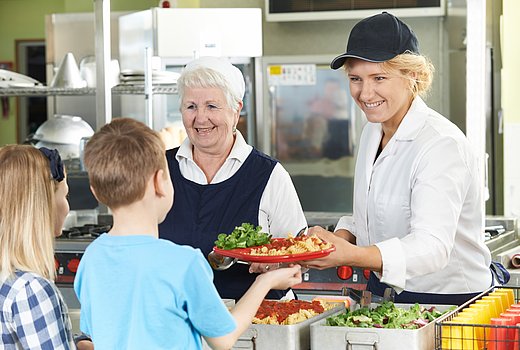 Zwei kinder stehen an der Ausgabetheke der Schulverfpelgung und erhalten Menü von freundlicher Servicekraft