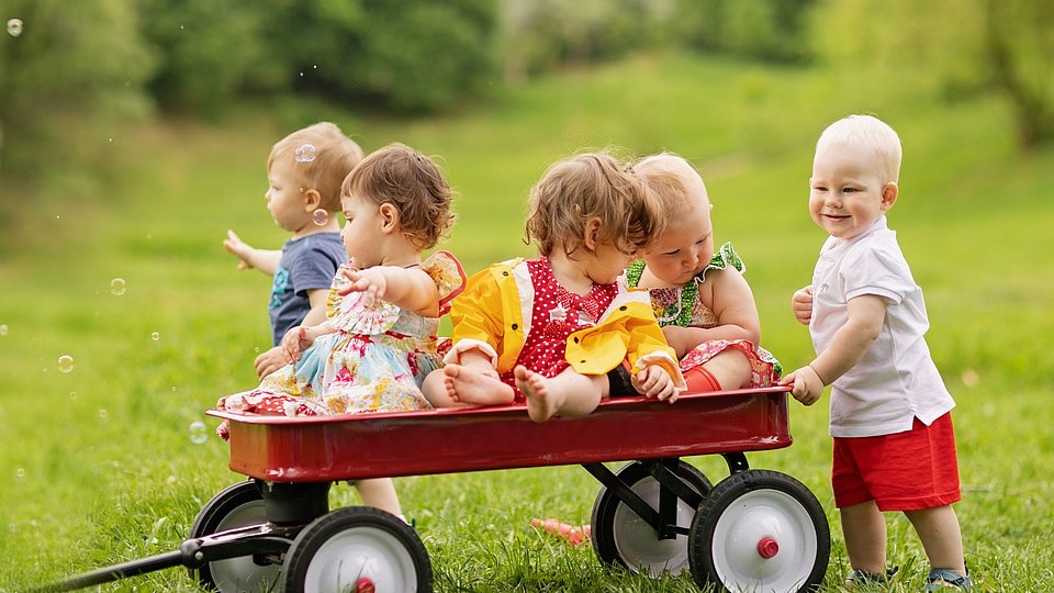 Eine Gruppe von fünf Kleinkindern sitzt und steht um einen roten Bollerwagen auf einer Wiese herum.