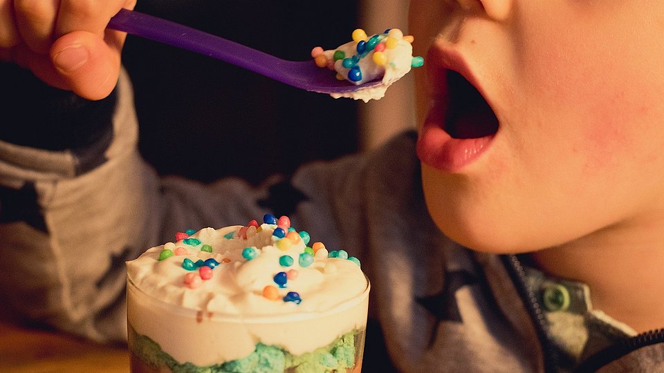 Das Foto zeigt in Großaufnahme, wie ein Kind einen Löffel zum geöffneten Mund führt. Der Löffel ist mit einer zuckerreich dekorierten Süßspeise gefüllt, vor dem Kind steht das Dessertglas.