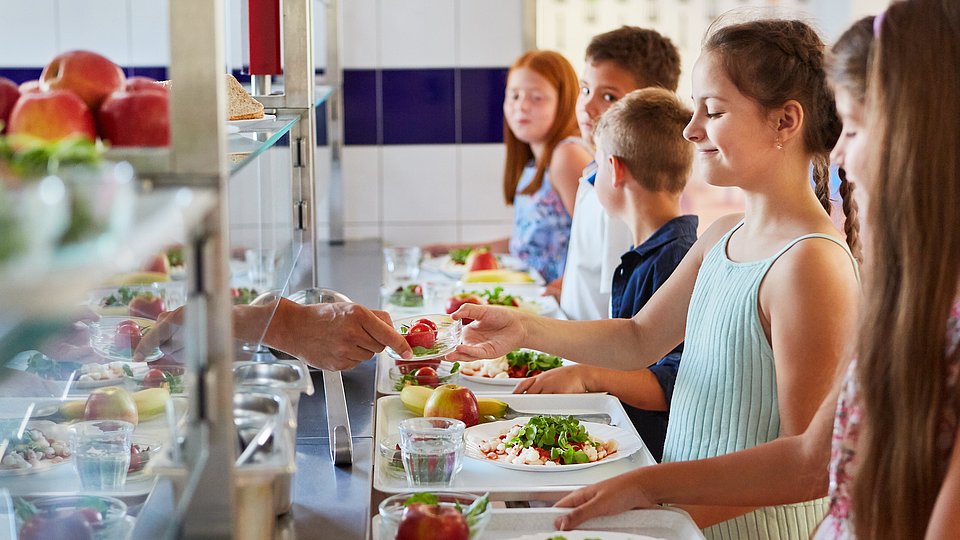 Kinder stehen in einer Schulmensa an der Ausgabetheke und suchen sich ihr Essen aus.