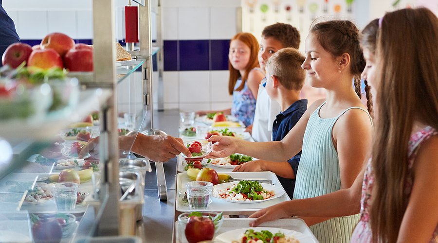 Kinder stehen in einer Schulmensa an der Ausgabetheke und suchen sich ihr Essen aus.