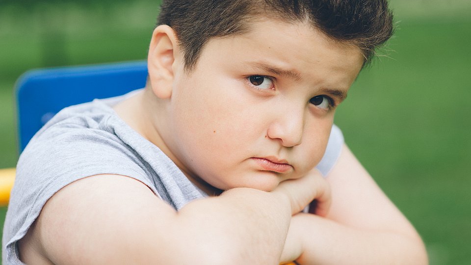 Ein etwas übergewichtiger Junge hat den Kopf auf seine Hände gestützt und schaut traurig in die Kamera. 