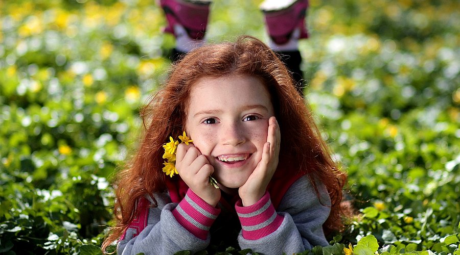 Kindergartenmädchen mit langen roten Haaren liegt bäuchlings auf einer Wiese und lacht mit Blumen in der Hand in die Kamera. 