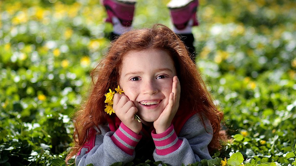 Kindergartenmädchen mit langen roten Haaren liegt bäuchlings auf einer Wiese und lacht mit Blumen in der Hand in die Kamera. 