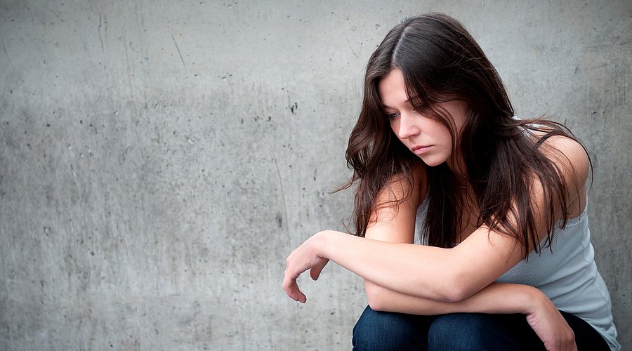 Jugendliche sitzt mit traurigem Gesicht vor grauer Wand.