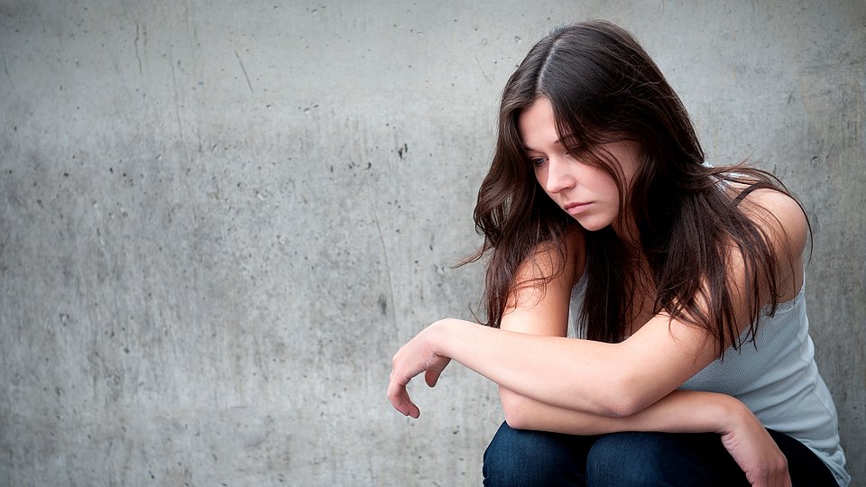 Jugendliche sitzt mit traurigem Gesicht vor grauer Wand.