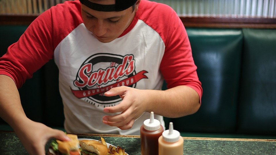 Jugendlicher Schüler mit Baseball-Cap sitzt an einem Tisch und isst einen Burger. 