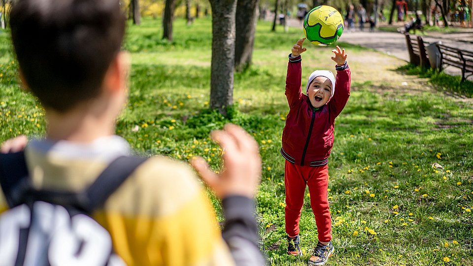 Ein Junge im Kitaalter wirft auf einem Rasen in einem Park einen gelben Ball einem älteren Jungen zu. Der kleinere Junge lacht fröhlich, der ältere Junge zeigt sich von der Rückenansicht, er trägt einen Rucksack und hält die Hände zum Fangen bereit. . 