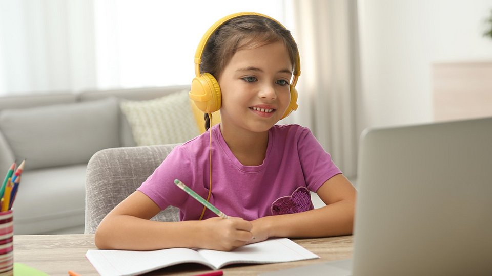 Ein Schulkind sitzt beim Home Schooling vor dem Laptop. Es hat gelbe Kopfhörer auf, vor ihm liegt ein geöffnetes Schulheft. 