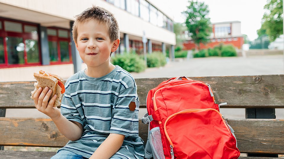 Ein Schuljunge mit kurzen blonden Stoppelhaaren sitzt verschmitzt lachend auf einer Holzbank vor seiner Schule und hält ein angebissenes Butterbrot in der Hand. Neben sich steht sein roter Schulrucksack.