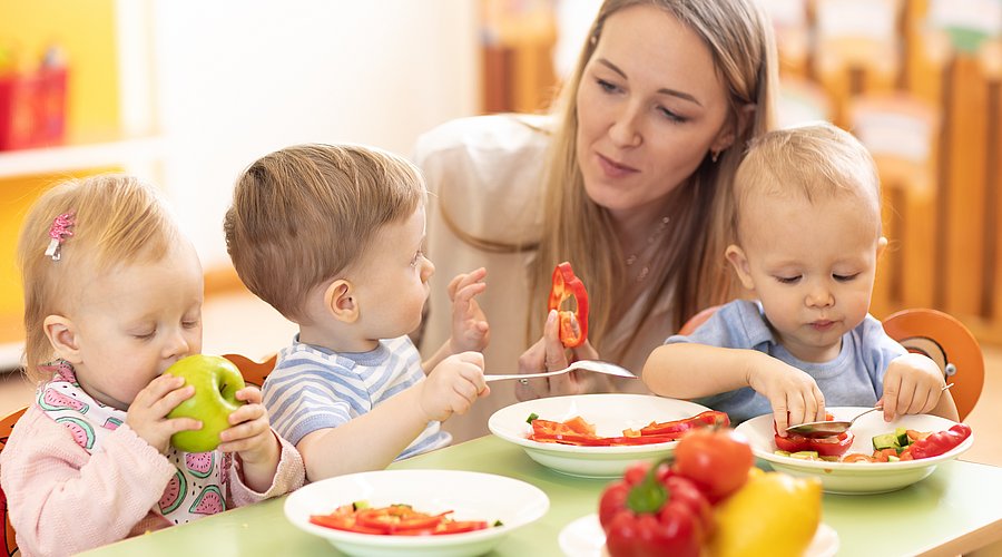 Kleine Kita-Kinder essen mit ihrer Erzieherin Obst- und Gemüserohkost.