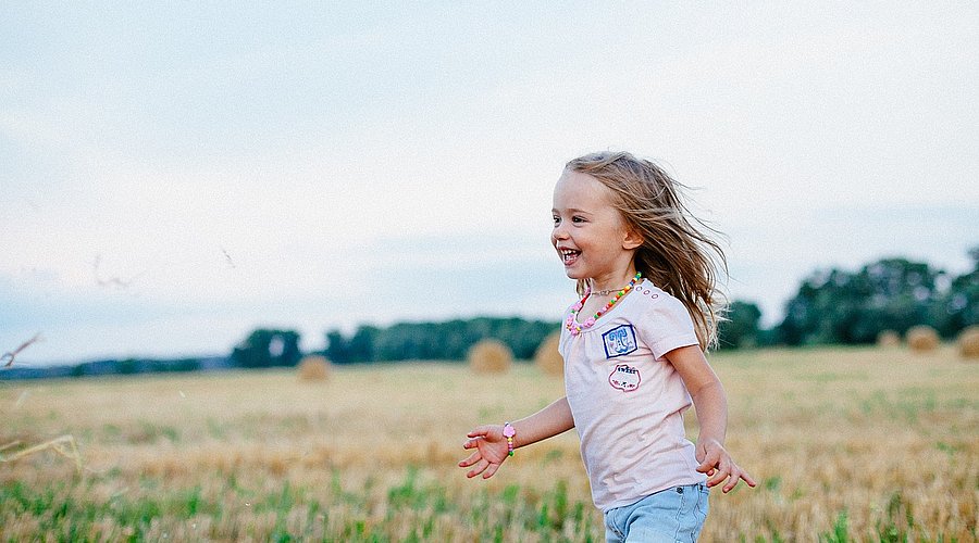 Kleines Mädchen mit langen blonden Haaren in T-Shirt und kurzer Jeanshose läuft am Rande eines sommerlichen Getreidefeldes.