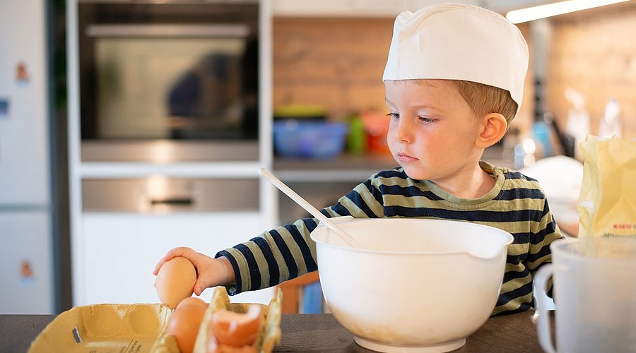 Ein Junge im Kindergartenalter steht an einem Küchentisch und hat eine Rührschüssel vor sich. Er nimmt ein rohes Ei aus einem Eierkarton. 