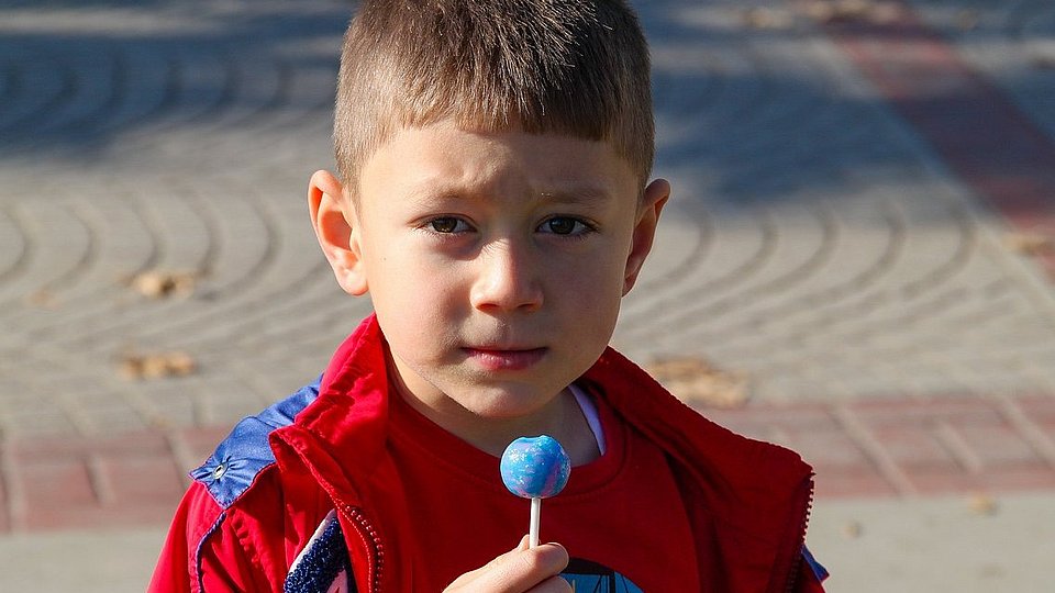 Ein kleiner Junge mit einer roten Jacke hält einen künstlich blauen Lolli in der Hand. 