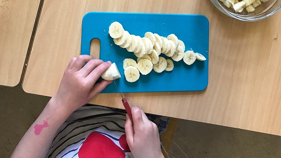 Kind schneidet in einer Kita eine Banane in Stücke. 