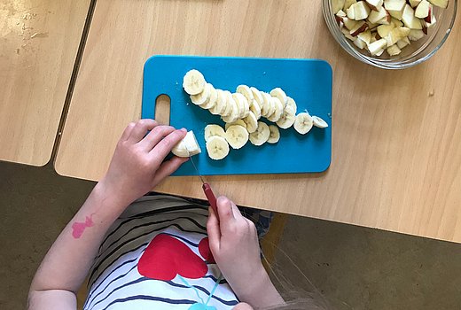 Kind schneidet in einer Kita eine Banane in Stücke. 