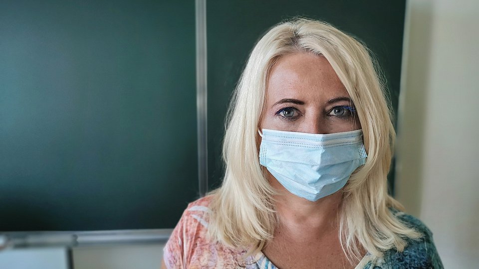 Eine junge Lehrerin mit Corona-Maske steht im Klassenraum vor einer leeren Tafel.