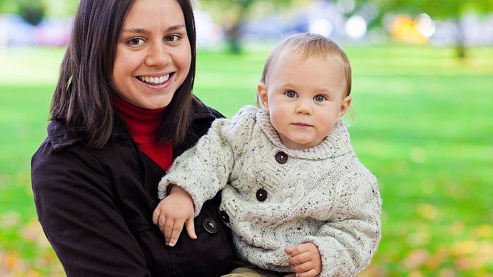 Eine junge Mutter steht in einem herbstlichen Park und hält ihr Baby auf dem Arm. 