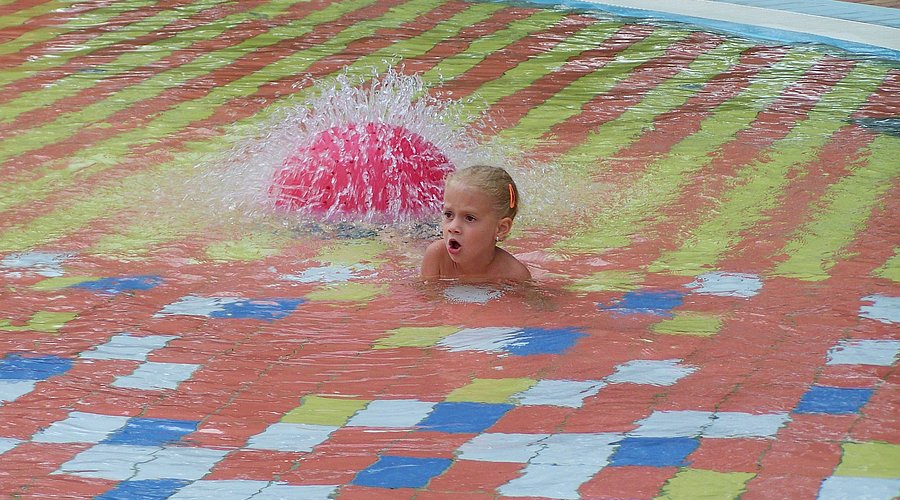 Kleines Mädchen sitzt in einem Kinderschwimmbad mit Wasserfontäne. 