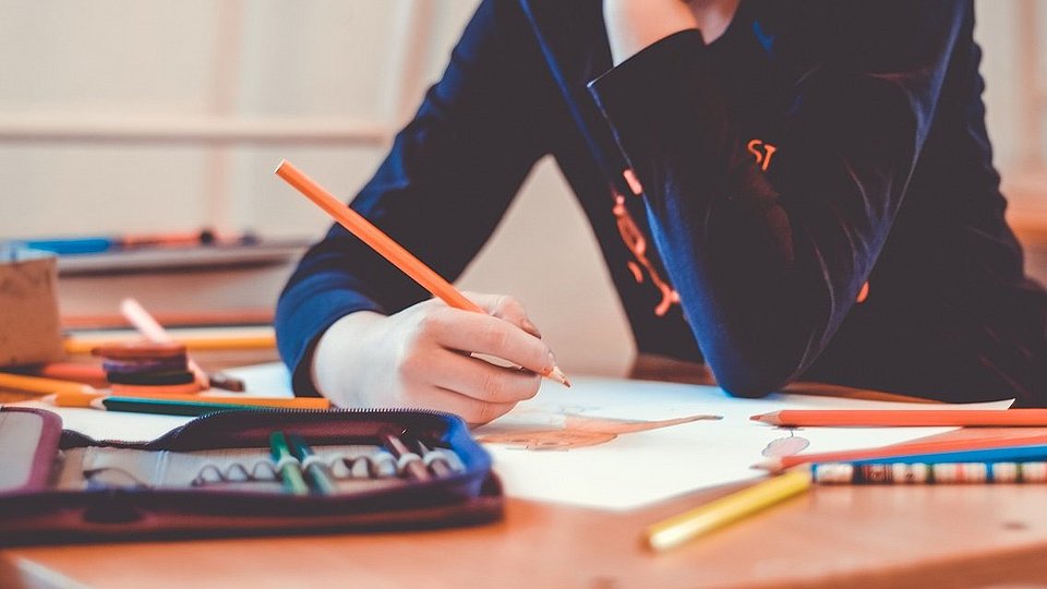 Jugendliche Schülerin sitzt am Schreibtisch, hat den Kopf in die Hände gestützt und schreibt in ein Schulbuch.