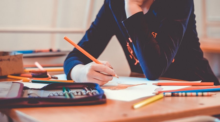 Jugendliche Schülerin sitzt am Schreibtisch, hat den Kopf in die Hände gestützt und schreibt in ein Schulbuch.