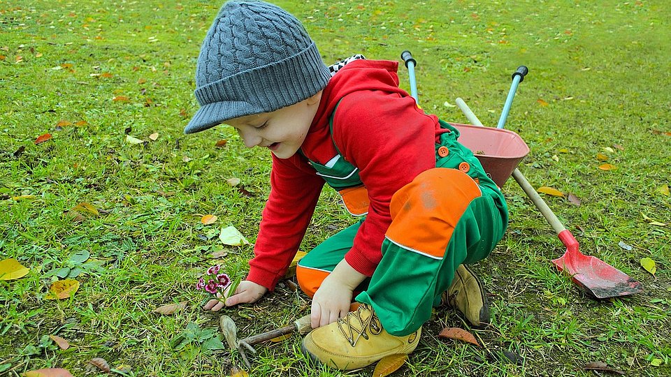 Ein Junge in Regenhose sitzt mit Gartenwerkzeug auf dem Rasen und pflanzt Blumen. 