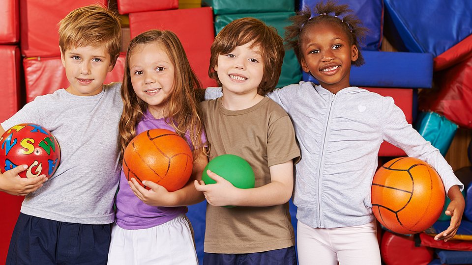 Vier Kinder mit unterschiedlicher Hautfarbe stehen untergehakt in einer Turnhalle vor bunten Sportmatten und halten jeweils einen Ball in der Hand. 