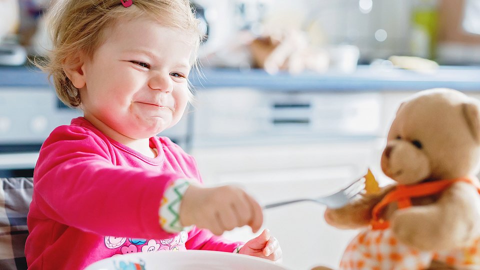 Kleines Mädchen sitzt im Hochstuhl am Tisch und füttert ihren Teddybären. 