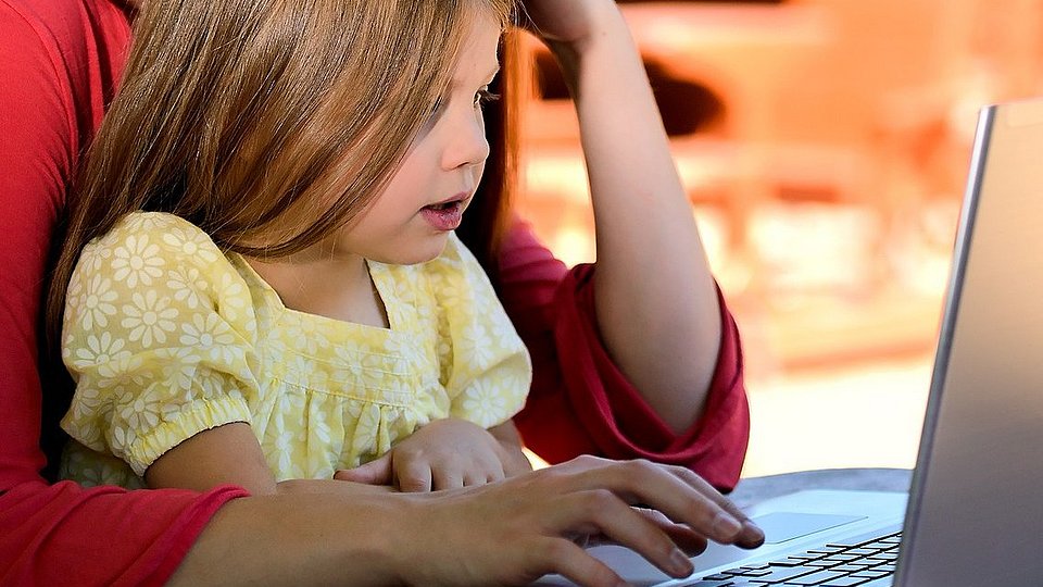 Eine Frau sitzt mit ihrer Tochter vor einem Laptop. Beide haben die Hände auf der Tastatur.