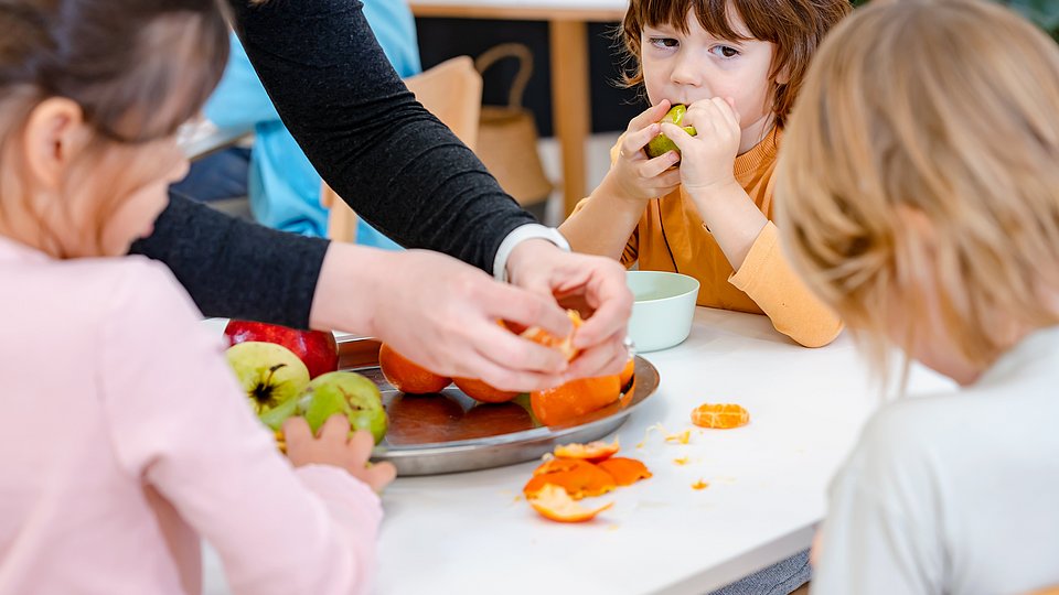 Kita-Kinder sitzen um einen Tisch und essen Obst von einer Obstplatte.