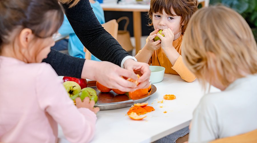 Kita-Kinder sitzen um einen Tisch und essen Obst von einer Obstplatte.